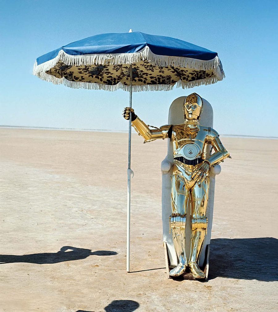 A gleaming humanoid robot lounges under a beach umbrella on an empty salt flat.